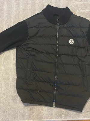Svart cardigan från Moncler - Svart cardigan från Moncler med quiltad front, ribbad krage och ärmar i mjukt material. Jackan har dragkedja framtill, två fickor med knapp och Moncler-logga på bröstet. Perfekt för kyliga dagar och snygg till streetwear.