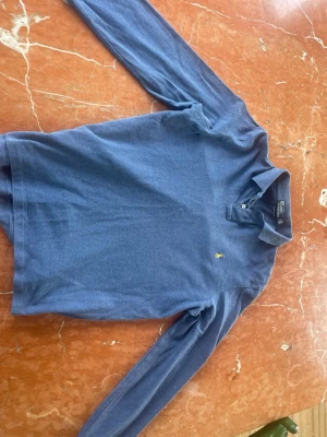Blå långärmad piké från Ralph lauren. - Klassisk blå långärmad henley från Polo sport med soprtig krage och två knappar framtill. Tillverkad i mjuk bomull och har den ikoniska lilla gula loggan broderad på bröstet. Perf lens för en avslappnad och stilren look.