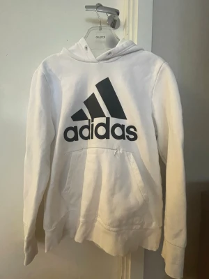 Vit hoodie från Adidas med logga - Säljer en vit hoodie från Adidas med stor svart logga på bröstet. Hoodien har huva med dragsko och en klassisk mag wrapping habitual ficka framtill. Perfekt för dig som gillar sportig och avslappnad stil.