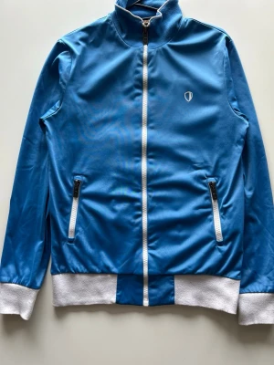 Ben Sherman blå träningsjacka, storlek S - Blå träningsjacka med hel dragkedja framtill och en hög krage. Jackan har vita muddar vid ärmslut och nederkant, samt vita detaljer längs dragkedjan och som en horisontell linje över ryggen.  📌 Detaljer Färg – blå Storlek – S Skick – bra   📏 Mått Axel till axel – 40 cm Axel till nederkant – 65 cm