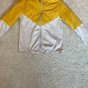 Gul och vit vindjacka med dragkedja - Snygg vindjacka i gult och vitt med dragkedja framtill och elastisk nederkant. Jackan har en cool färgblock-design och är tillverkad i ett lätt, vindtätt material. Perfekt för blåsiga dagar och enkel att matcha med olika outfits. Strl 152. Använd flertal gånger. OBS den har en fläck på baksidan, se bild. 