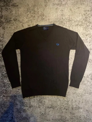 Svart V-ringad tröja Fred Perry - Klassisk svart stickad tröja från Fred Perry med V-ringning och blått broderat logotypmärke på bröstet. Tröjan har långa ärmar och ribbade muddar vid ärmslut och nederkant. Perfekt till lager-på-lager och enkel att matcha.