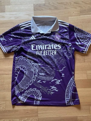 Lila Real Madrid matchtröja drake - Säljer en lila Real Madrid matchtröja från Adidas med vit krage och detaljer. Tröjan har ett stort drakmönster i silver, klubbmärke på bröstet och Emirates Fly Better-tryck på framsidan. Perfekt för fotboll eller streetwear. Det står storlek XL men passar bättre som storlek L
