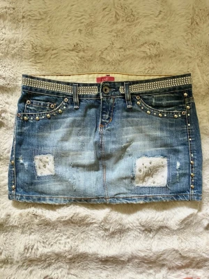 Kort jeanskjol med nitar och patchar - Säljer en blå kort jeanskjol med coola slitningar, patchar och nitar längs fickorna. Kjolen har en glittrig bälteffekt upptill och klassiska fickor både fram och bak. Perfekt för dig som gillar en edgy och trendig stil. Storlek M men passar snarare S enligt mig! Dubbelkolla bilderna med mått samt på mig för referens!!! 🩷