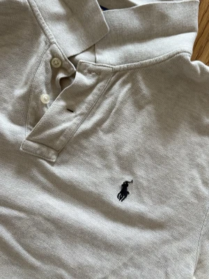 Grå pikétröja från Ralph Lauren - Klassisk beige pikétröja från Ralph Lauren med korta ärmar och broderad logga på bröstet. Tröjan har krage, knappar vid halsen och är tillverkad i mjuk bomull. Perfekt för en avslappnad och stilren look.