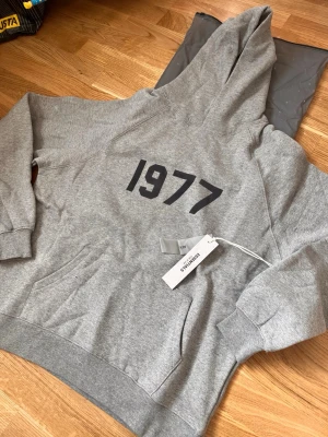 Grå Essentials Fear of God hoodie - Snygg grå hoodie från Essentials Fear of God med svart tryck '1977' på bröstet. Klassisk passform med huva och mjukt material som känns skönt mot huden. Perfekt för dig som gillar streetwear och vill ha en stilren look.