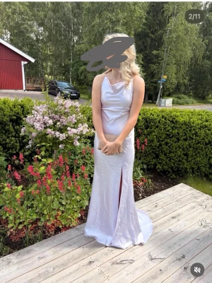 Ljuslila långklänning med slits - Elegant ljuslila långklänning i ett glansigt material med slits på sidan och mjukt draperad urringning. Klänningen är ärmlös och har en smickrande passform som följer kroppen. Perfekt för speciella tillfällen. ny pris ca 1300kr