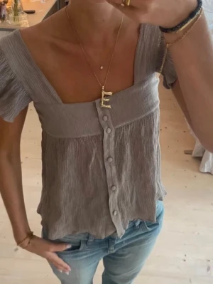 Beige blus med knappar och kort ärm - Säljer en luftig beige blus med korta ärmar och knappar framtill. Toppen har en fyrkantig ringning och är tillverkad i ett tunt, skrynkligt tyg som ger en avslappnad vibe. Perfekt till jeans eller kjol för en chill look.
