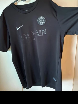 Svart PSG x Balmain t-shirt Nike - Snygg svart t-shirt från Nike i samarbete med PSG och Balmain. T-shirten har korta ärmar, rund halsringning och tryck med Paris Saint-Germain-logga samt Balmain Paris på bröstet. Tillverkad i ett mjukt material som känns skönt mot huden. 