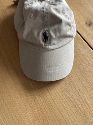 Beige keps från Polo Ralph Lauren - Snygg beige keps från Polo Ralph Lauren med klassisk mörkblå broderad logga framtill. Kepsen är tillverkad i 100% bomull och har en böjd skärm. Perfekt accessoar för en avslappnad och stilren look.