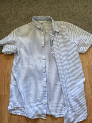 Blårandig kortärmad skjorta - Snygg kortärmad skjorta med blå och vita ränder. Klassisk krage och knappar framtill. Perfekt till sommaren och lätt att matcha med jeans eller shorts. Tillverkad i mjuk bomull för en skön känsla.