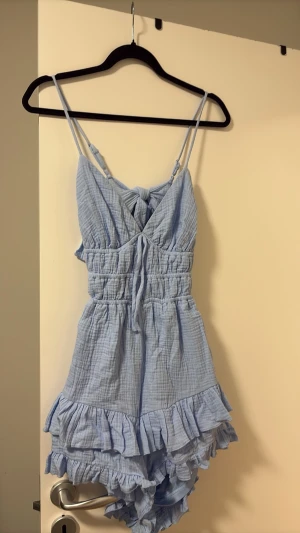 Ljusblå volangjumpsuit med smala axelband - Supersöt ljusblå jumpsuit med smala justerbara axelband, knytdetalj framtill och volanger längs shortsen. Tillverkad i ett mjukt, krinklat bomullstyg som känns luftigt och bekvämt. Perfekt för varma dagar när du vill ha något lätt och stilrent.