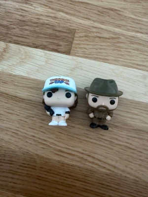 Two  Funko Pops, hopper and Dustin from stranger things - Säljer två Funko Pop-figurer i vinyl. Ena figuren har en blå keps med texten 'Thinking Cap', vit tröja och bruna detaljer. Den andra figuren har grön hatt, brunt skägg och gröna kläder. Perfekta för samlare eller som dekoration.