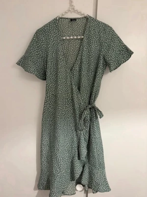 Grön prickig omlottklänning Vero Moda - Snygg grön omlottklänning från Vero Moda med vita prickar och volangkant nertill. Klänningen har korta ärmar och knyt i midjan för en smickrande passform. Perfekt för vår och sommar!