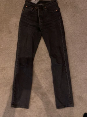 Svarta raka jeans från Levi's - Säljer ett par svarta raka jeans från Levi's med klassisk femficksdesign och hög midja.  Perfekta för dig som gillar en clean och tidlös stil. Hål på vänster baksida, men har lagat det så det syns knappt nåt när man har de på sig! 