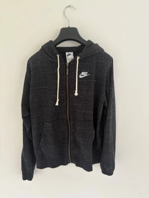 Svart zip-hoodie från Nike - Mysig svartmelerad zip-hoodie från Nike med vit logga på bröstet. Tröjan har huva med vita dragsnören, fickor framtill och ribbade muddar vid ärmslut och nederkant. Perfekt för en avslappnad stil. (Sportswear Gym Vintage Women’s Full-Zip Hoodie Black/White. Nypris 999 kr. Aldrig använd men prislappen borttagen. 