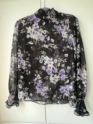 Blommig transparent blus med volang - Snygg svart transparent blus med lila och vita blommor. Hög krage med smock, långa ärmar och breda smockade muddar med volangkant. Tunn och luftig polyester, snyggt till ex vita jeans i sommar👌 