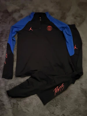PSG tracksuit träningsset från Jordan, svart/blå - Snyggt PSG träningsset från Jordan med långärmad svart tröja och blå ärmar, röda loggor på bröstet och ärmen samt texten 'Paris' på byxan. Perfekt för fotbollsträning eller chill. Materialet är mjukt och stretchigt för bästa komfort.
