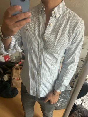 Randig skjorta med knappkrage - Säljer en snygg ljusblå och vit randig skjorta från Polo Ralph Lauren med klassisk knappkrage och broderad logga på bröstet. Skjortan har långärmad design, knappar framtill och är tillverkad i bomull. Perfekt för dig som gillar stilrena och tidlösa plagg. En liten fläck går nog att få bort