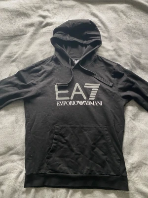 Svart EA7 Emporio Armani hoodie - Snygg svart hoodie från EA7 Emporio Armani med stor logga framtill. Klassisk huva med dragsko och en stor magficka. Tillverkad i mjukt material som känns skönt mot huden. Perfekt för dig som gillar stilrena och exklusiva plagg.