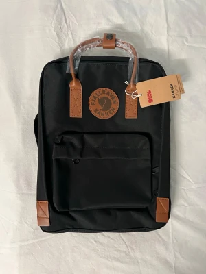 Fjällräven ryggsäck - Stilren svart Kånken ryggsäck från Fjällräven med bruna läderdetaljer på handtag och hörn. Klassisk fyrkantig form, frontficka med dragkedja och justerbara axelband. Materialet är slitstark canvas och läder, perfekt för att bära dina grejer till skolan eller stan. Ryggäcken är för liten för min dator och säljs på grund av det. 1: 1. Hade kunnat tänka mig att gå ner lite i pris vid snabb och smidig affär. Vid någora frågor är det bara att skriva 