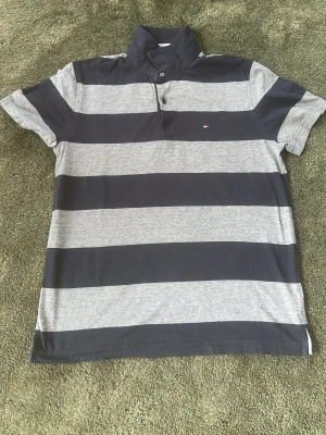 Randig pikétröja från Tommy Hilfiger - Klassisk pikétröja från Tommy Hilfiger med breda marinblå och grå ränder. Tröjan har korta ärmar, krage och knappar framtill. Tillverkad i mjuk bomull och har den ikoniska lilla loggan broderad på bröstet.