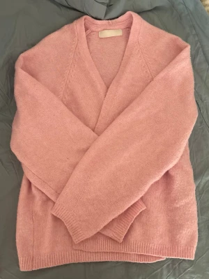 Ljusrosa stickad kofta - Mysig ljusrosa stickad kofta med öppen front och långa ärmar. Koftan har en avslappnad passform och ribbade muddar vid ärmslut och nederkant. Perfekt att slänga över en t-shirt eller topp för extra värme.