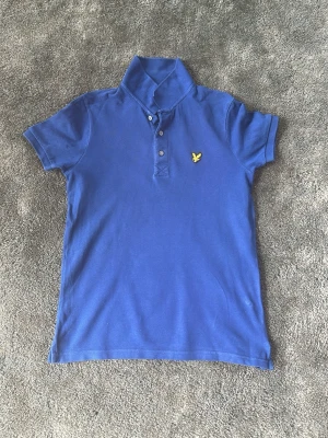 Blå pikétröja från Lyle & Scott - Säljer en blå pikétröja från Lyle & Scott med klassisk krage och tre knappar framtill. Tröjan har ett gult fågelbroderi på bröstet och är tillverkad i mjuk bomull. Perfekt för dig som gillar stilrena och sportiga plagg.