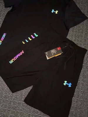 Under armour short  - Snyggt svart t-shirt set från Under Armour med färgglada loggor och textdetaljer i regnbågsfärger. Tillverkad i mjukt och funktionellt material som passar perfekt till träning eller sport. Modern passform och cool design för dig som gillar sportiga kläder.