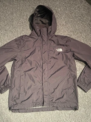 Mörkgrå regnjacka från The North Face St:S - Använt fåtal gånger. Snygg svart regnjacka från The North Face med huva och dragkedja framtill. Jackan har elastiska muddar vid ärmsluten, två sidofickor med dragkedja och logga på bröstet. Tillverkad i lätt och vattenavvisande material, perfekt för regniga dagar.