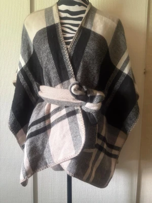 Rutig ponchosjal i svart och beige - Snygg och stor sjal i ponchomodell med rutigt mönster i svart, beige och grått. Sjalen har ett bälte i midjan som knyts för en cool och avslappnad look. Perfekt att slänga över axlarna för extra stil. Materialet känns mjukt och värmande.