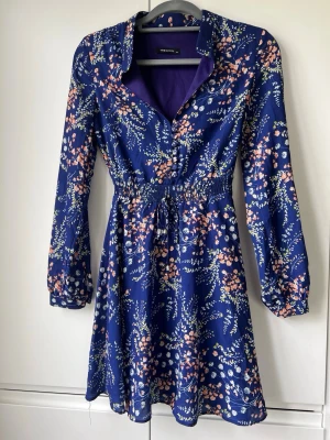 Blommig blå klänning från Trendyol - Supersöt blå klänning från Trendyol med blommigt mönster i vitt och rosa. Klänningen har lång ärm, markerad midja med resår och knappar och knytband. Fodrad. 