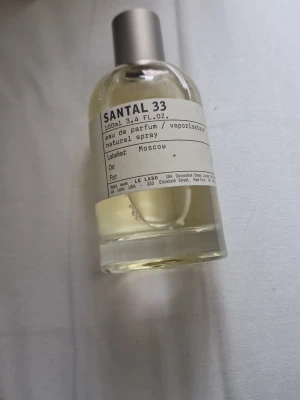 Le Labo Santal 33 Eau de Parfum 100ml - Le Labo Santal 33 Eau de Parfum i en stilren, transparent glasflaska med metallock. Doften är ikonisk och unisex, med träiga och kryddiga toner. Flaskan rymmer 100 ml och har en minimalistisk etikett med svart text.