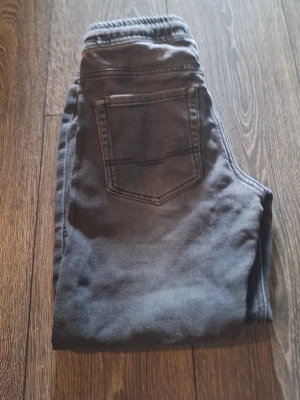 Grå jeans från Pepperts med resår - Säljer ett par grå jeans från Pepperts med resår i midjan och klassisk bakficka. Jeansen har en avslappnad passform och är perfekta för en chill och bekväm stil. Materialet är mjukt och stretchigt för extra komfort.