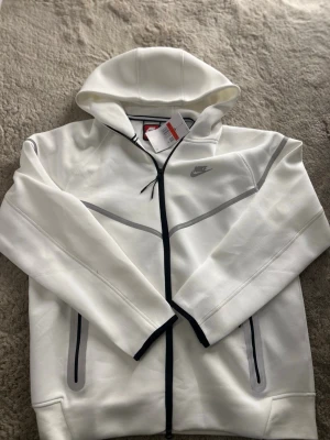 Vit Nike hoodie med dragkedja - Snygg vit hoodie från Nike med hel dragkedja och gråa detaljer. Hoodien har två fickor med dragkedja och en klassisk Nike-logga på bröstet. Tillverkad i mjukt material och har en huva för extra stilpoäng. Perfekt för en sportig och avslappnad look.