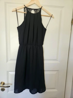 Klänning  Gina Tricot 34 - Svart klänning med tunna axelband och snygg spetsdetalj i ryggen. Klänningen har veckad front, markerad midja och faller i en mjuk, rak siluett. Perfekt för dig som gillar stilrena plagg med en twist.