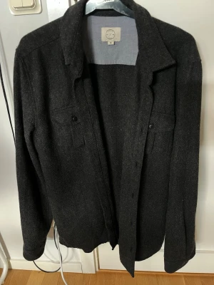 Mörkgrå overshirt - Stilren mörkgrå overshirt från Suit Apparel i storlek M. Skjortjackan har klassisk krage, två bröstfickor med knappar och är tillverkad i ett tjockare, mjukt tyg som ger en exklusiv känsla. Perfekt lager-på-lager-plagg.