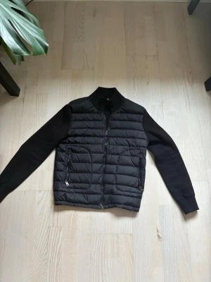 Svart Moncler cardigan - Svart cardigan från Moncler. Den har dragkedja framtill, två fickor med dragkedja och Moncler-logga på ärmen. Snygg och stilren modell som funkar till mycket.