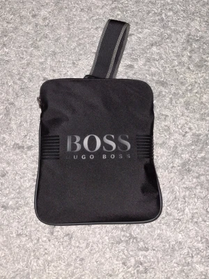 Svart axelväska från Hugo Boss - Snygg svart axelväska från Hugo Boss med justerbar axelrem och stort BOSS-tryck på framsidan. Väskan har en fyrkantig form och är tillverkad i slitstarkt tygmaterial. Perfekt för att bära det viktigaste och ger en clean och modern look.