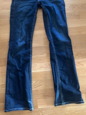 Mörkblå bootcut jeans med kontrastsömmar - Snygga mörkblå bootcut jeans med vita kontrastsömmar och klassiska bakfickor med lock och knapp. Storlek 170. Jeansen har en utsvängd passform nedtill och är tillverkade i denim. Perfekta för en avslappnad och trendig look.