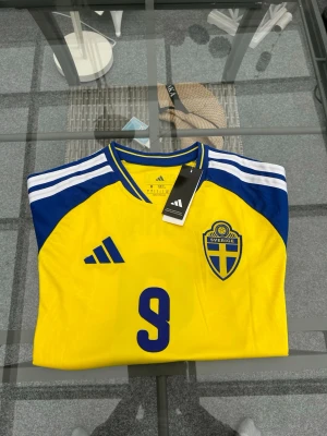 Ny! Sverige-tröja Isak 9 (Adidas) 🇸🇪 - Säljer denna snygga och helt oanvända Sverige-tröja med Isak #9 på ryggen! ⚽️💙💛 Den har aldrig använts och alla lappar sitter kvar. Säljer den enbart för att jag ångrade köpet och vill att någon annan ska få glädje av den istället. Perfekt passform (Slim fit) och superfräsch. Info: ✨ Märke: Adidas ✨ Storlek: M ✨ Skick: Helt ny med tags (NWT) Skriv ett DM vid frågor eller om du vill ha fler bilder! Kan skickas spårbart. 📦 