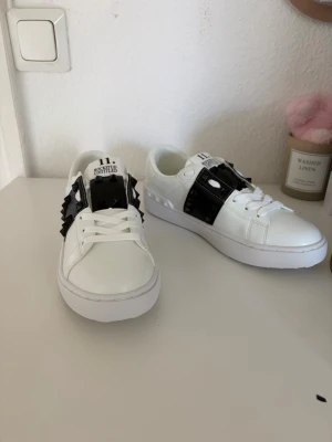 Valentino Rockstud Untitled sneakers - Snygga vita sneakers från Valentino med svart band över vristen dekorerat med ikoniska nitar. Skorna har en chunky sula och klassisk låg profil. Tillverkade i slätt läder med coola detaljer som ger en edgy vibe.