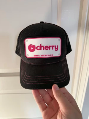 Cherry x John Hatter keps - Snygg svart keps med rosa sömmar och ett stort vitt märke framtill med texten 'cherry' i rosa. Kepsen har mesh på sidorna och bak för extra ventilation och justerbar passform. Perfekt för dig som vill sticka ut med en unik accessoar.