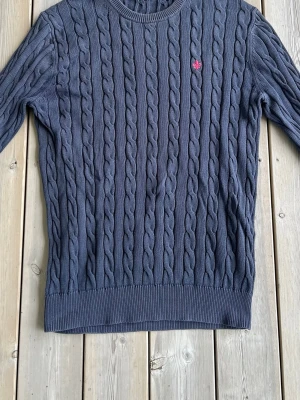 Mörkblå kabelstickad tröja - Kabelstickad tröja i mörkblått med rund hals och ribbade muddar. Tröjan har ett diskret rött broderat emblem på bröstet och är tillverkad i mjukt material. Föredetta hål i halsen men som är sytt ihop. Perfekt för dig som gillar klassisk och stilren look.
