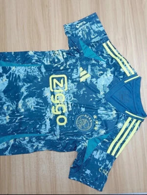 Ajax blå matchtröja Adidas - Ajax matchtröja i blått med gult mönster och gula detaljer. Adidas-logga och Ziggo sponsortryck på bröstet samt Ajax klubbmärke med tre stjärnor. Korta ärmar och rund hals. Tillverkad i lätt och ventilerande material, perfekt för fotbollsträning.