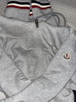 Grå kofta från Moncler - Säljer ett par grå kofta från Moncler med klassiska ränder i rött, vitt och blått vid bensluten. Koftan har en avslappnad passform och är tillverkade i mjukt bomullsmaterial. Perfekta för chill dagar eller när du vill vara bekväm.