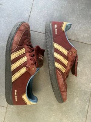 Adidas Wales Bonner vinröda sneakers - Säljer ett par Adidas Wales Bonner sneakers i vinrött med klassiska gula ränder på sidorna. Skorna har svart sula, blått innerfoder och gulddetaljer vid snörningen. Tungan har Adidas-logga och modellen är retroinspirerad.