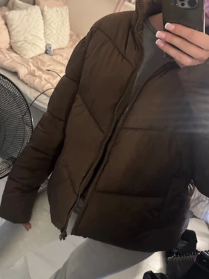 Brun pufferjacka från ASOS DESIGN - Snygg brun pufferjacka från ASOS DESIGN med hög krage och dragkedja framtill. Jackan har en boxig passform och breda kanaler som ger en modern look. Tillverkad i ett mjukt, vadderat material som håller dig varm under kalla dagar.