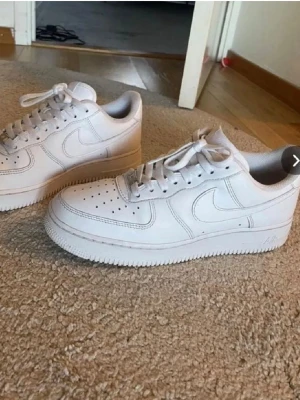 Nike Air Force 1 vita sneakers - Klassiska vita Nike Air Force 1 sneakers i lågt utförande. Skorna har en clean design med perforerad tå, vita skosnören och den ikoniska swoosh-loggan på sidan. Tillverkade i läder med en rejäl sula för skön komfort.