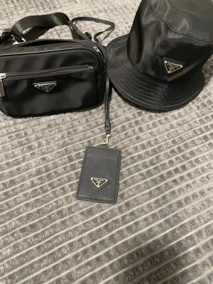 Svart bucket hat från  - Snygg svart bucket hat från  i glansigt nylonmaterial med den klassiska triangulära logotypen framtill. Hatten har en mjuk, rundad form och bred brätte som ger en cool streetwear-vibe. Perfekt accessoar för dig som vill sticka ut med exklusiv stil.